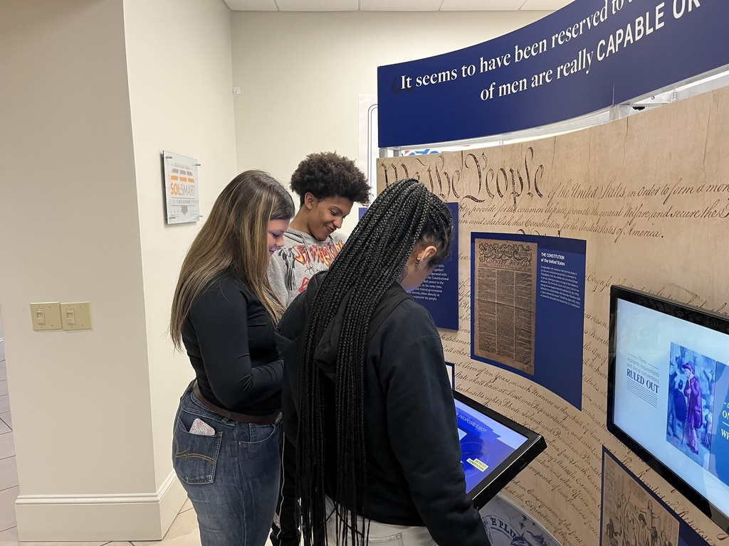 Three people look at a display, with one reading from a tablet. A large screen and text are visible.