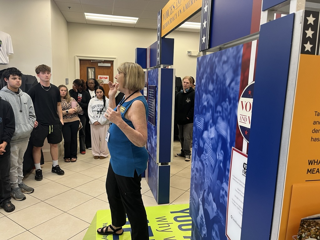 People gather around a woman in a blue top and black pants speaking in a room with a blue and orange wall.
