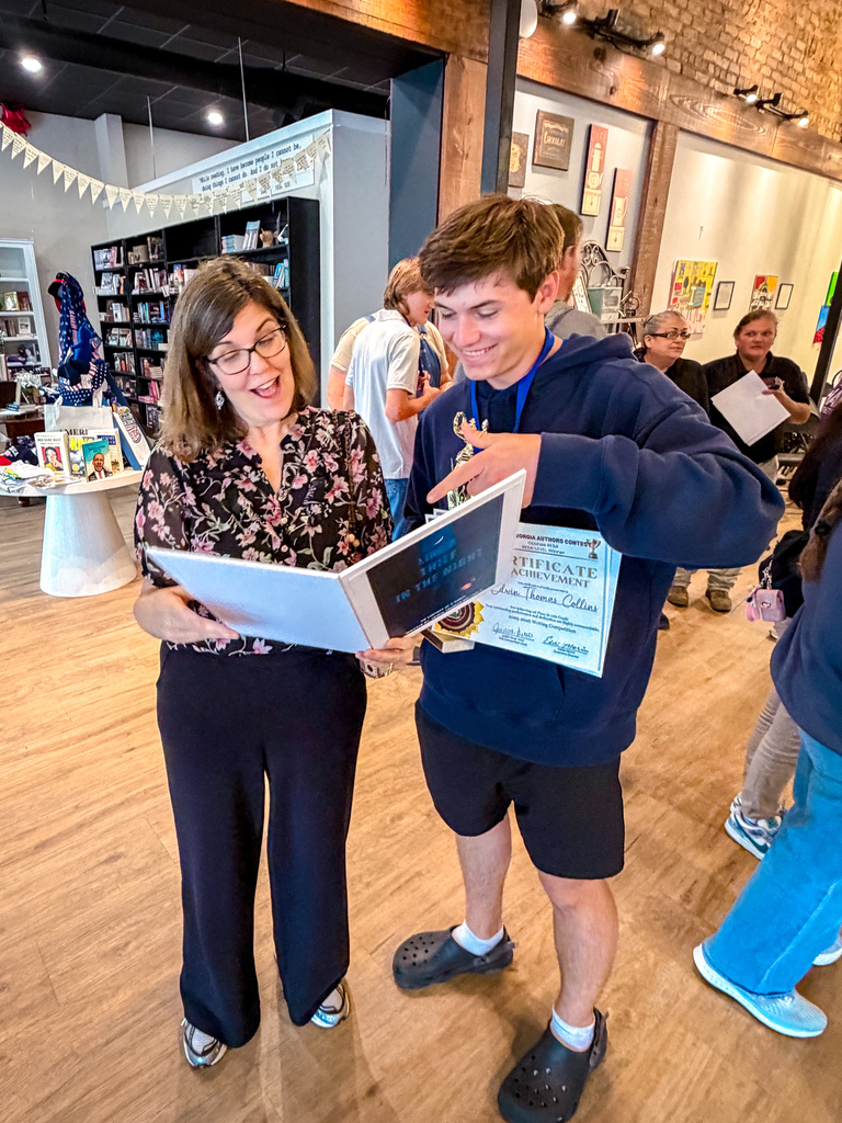 Woman with glasses holds certificate as young man points to it. They stand in a room with wooden floors and people around them.