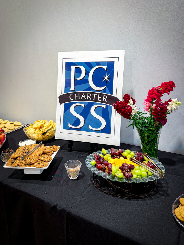 A table with a blue sign reading "PC Charter SS" displays various foods, including cookies, fruits, and salad.