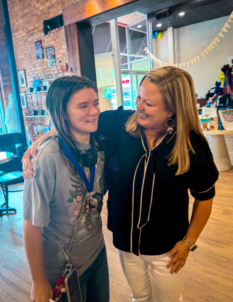 Two women, one with a medal around her neck, standing together in a room with wooden floors and an open door.