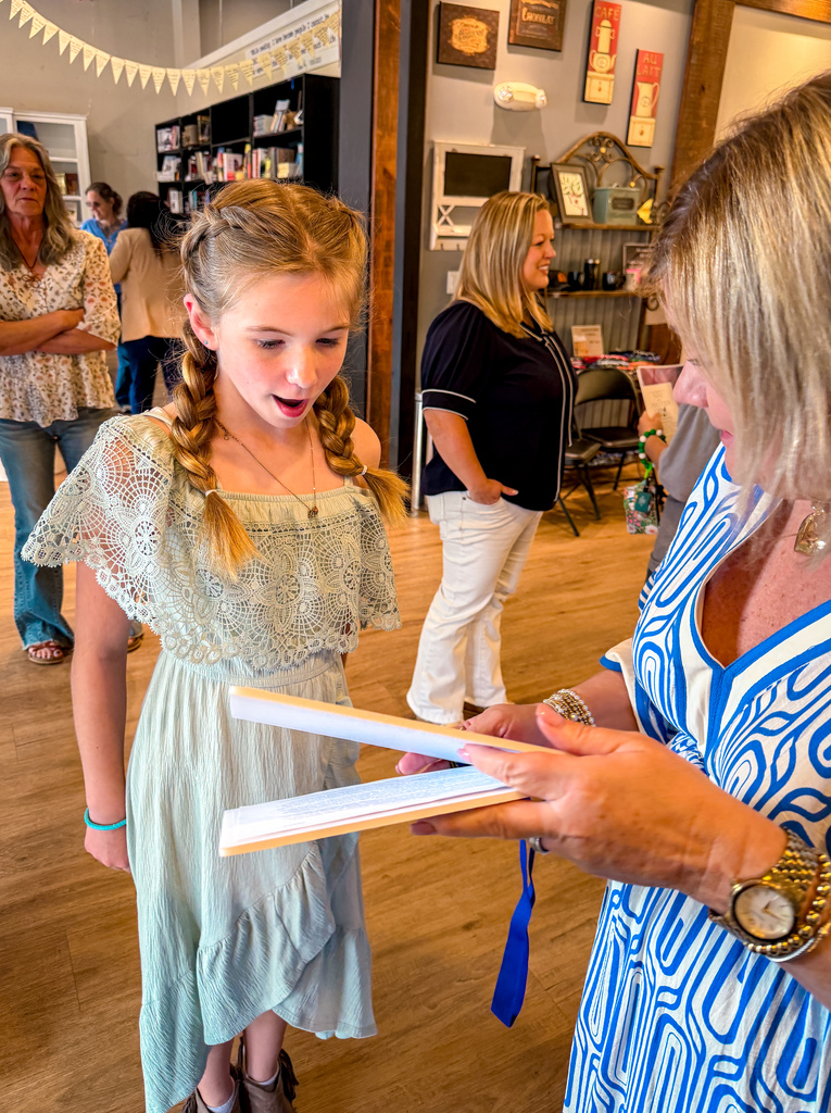 A young girl with braided hair in a pale green dress stands with an adult woman. They hold papers.