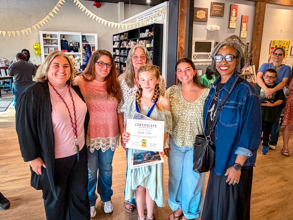 Five women and a young girl stand close together, smiling, and posing for a photo. One girl holds a certificate.