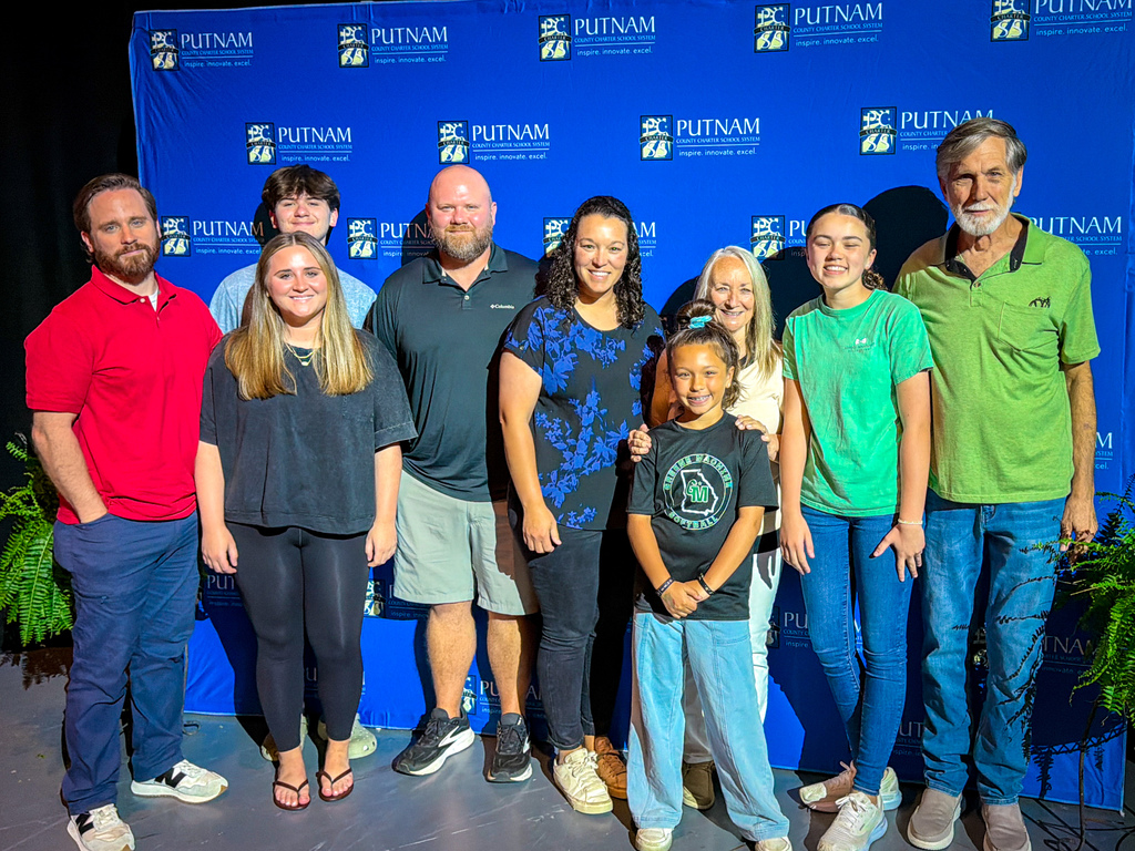 Nine people stand together in front of a blue banner, smiling for a photo. They wear casual clothing.