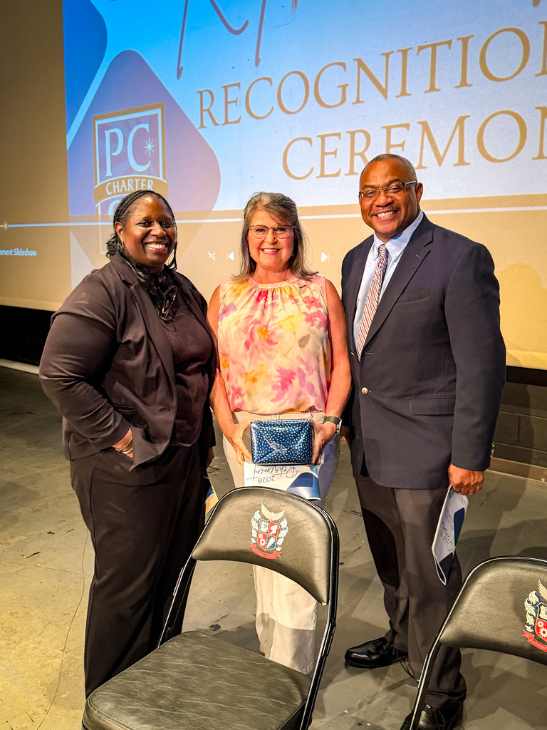 Three people stand together. The person in the middle holds an award. Behind them, a sign reads "Recognition Ceremony."