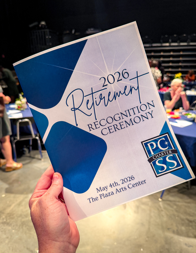 A hand holds a blue and white event program for a 2026 Retirement Recognition Ceremony. Background shows a room with tables and chairs.