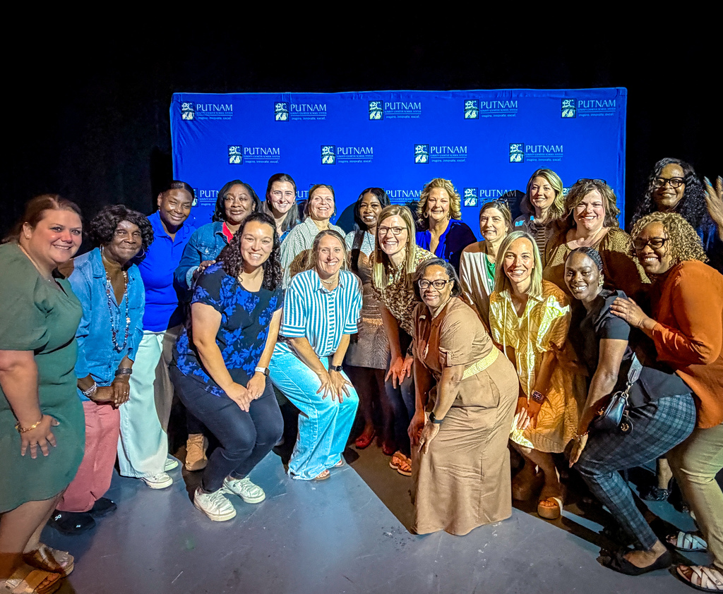 A group of women stands on a stage, smiling, with a blue backdrop displaying logos. They wear varied clothing and some pose with their arms raised.