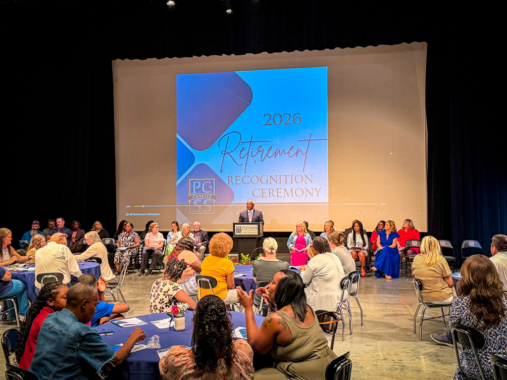 An indoor event showing a crowd of people seated around tables. A man speaks from a podium. A projector screen displays "2026 Retirement Recognition Ceremony".