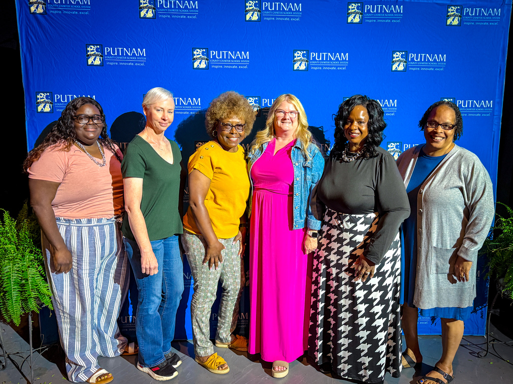 Six women stand in front of a blue backdrop with logos. Each wears different clothing and footwear, including sandals.