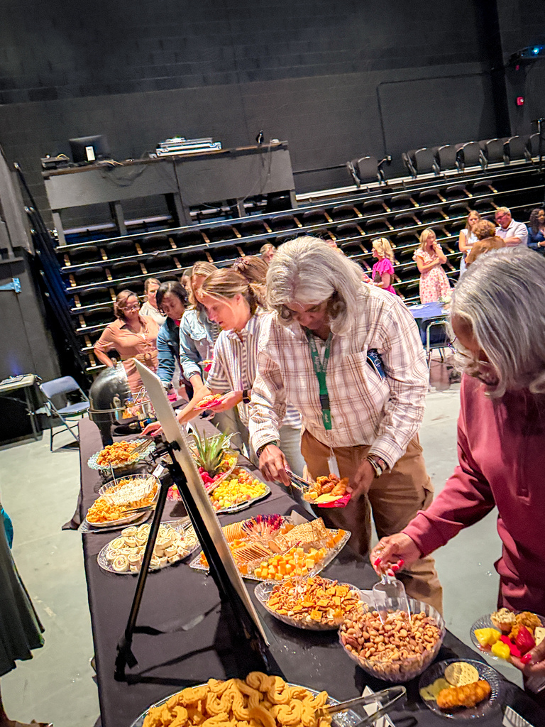 Several people gather around a table with a variety of foods on it. An older woman in a maroon shirt looks at the food.