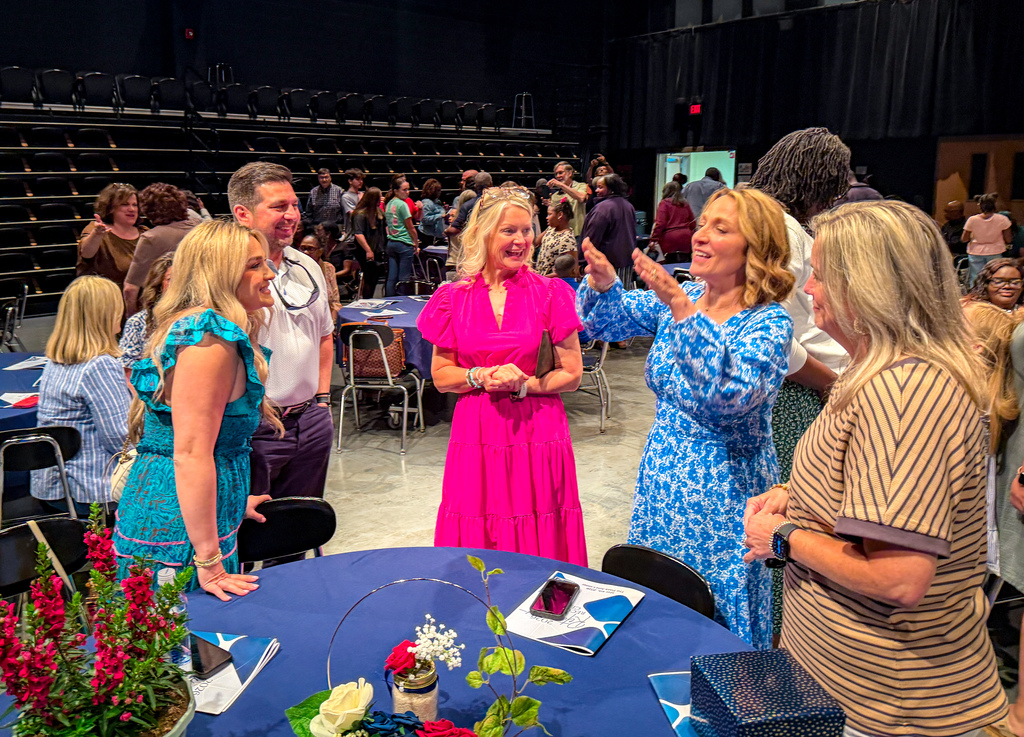 People in a large room with empty seats, a table with flowers, and a group of women talking.