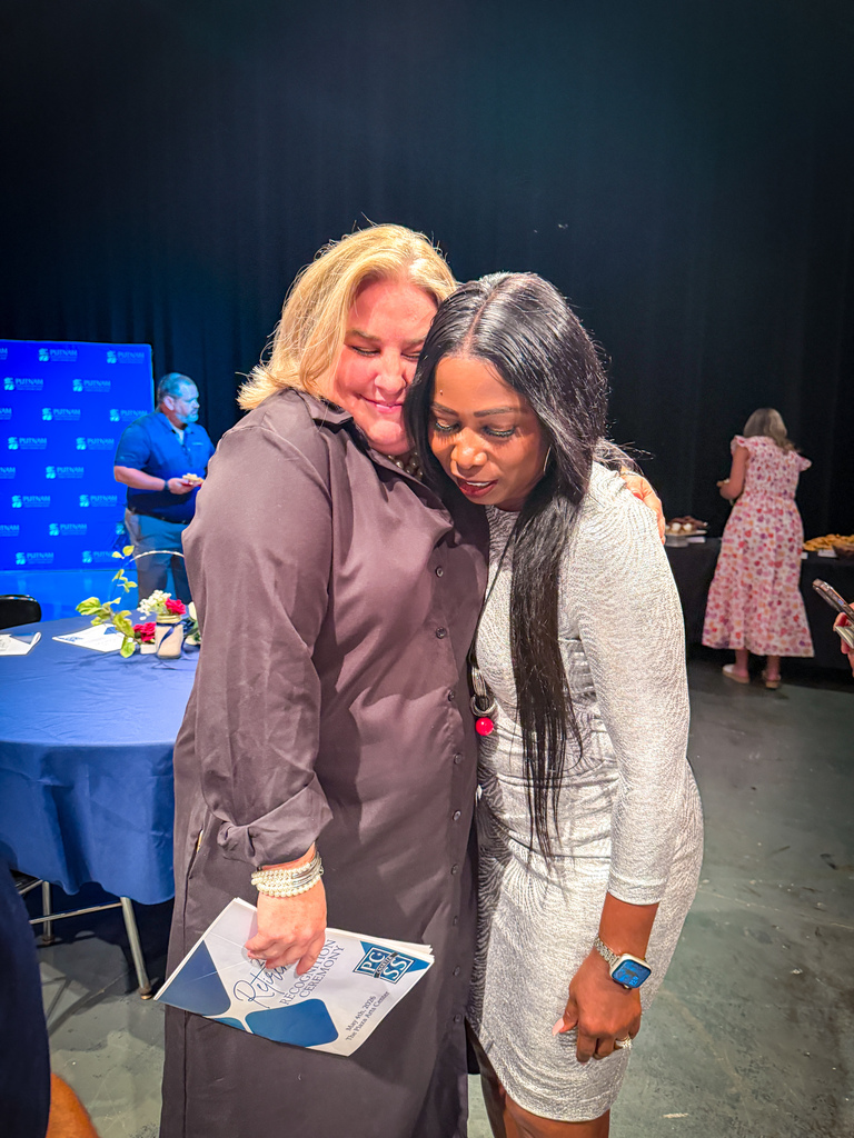 Two women hugging, one holding a booklet. They are indoors with a blue table, flowers, and a large black backdrop.