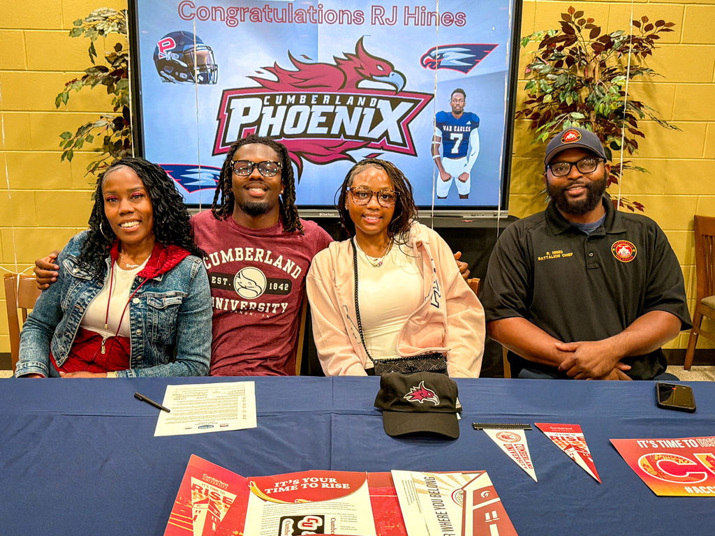 Four people in casual attire seated at a table with promotional items, behind them a large screen with text and a football player.