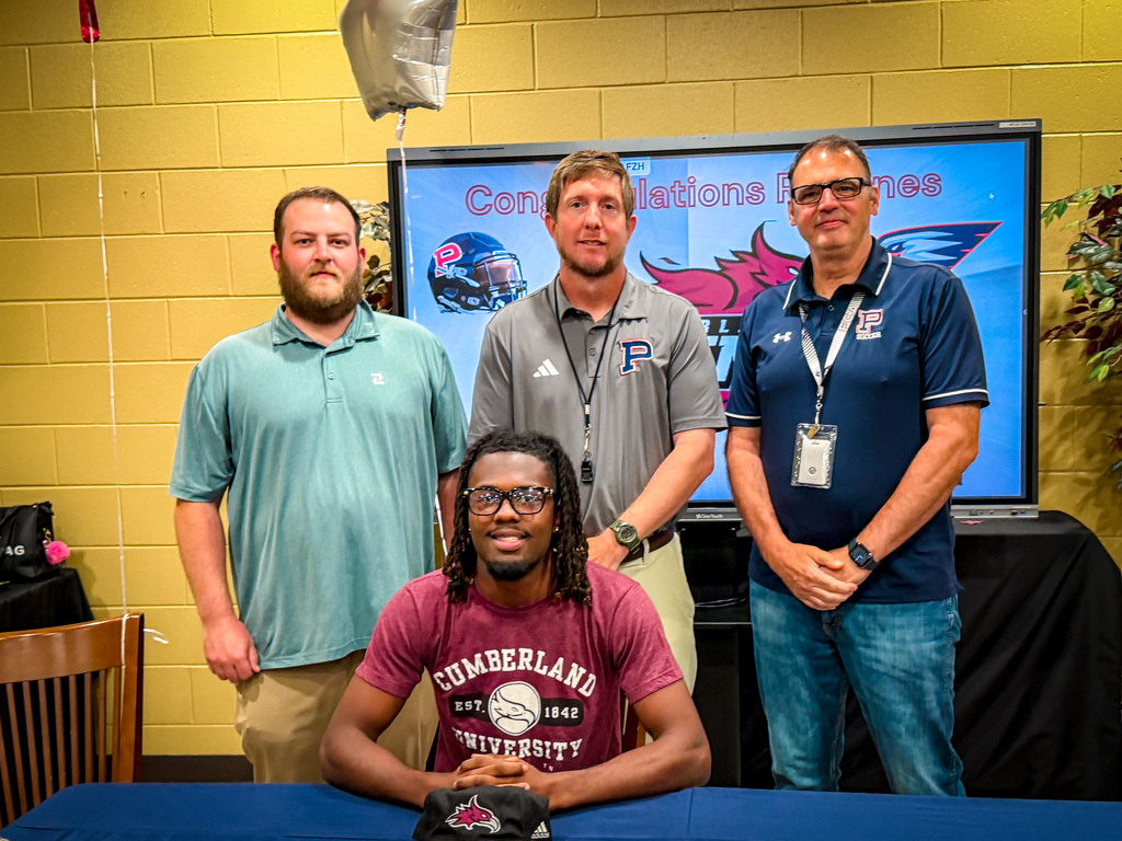 Four men standing behind a table; one man is seated, wearing glasses and a watch. A television screen behind them displays a team logo and text.