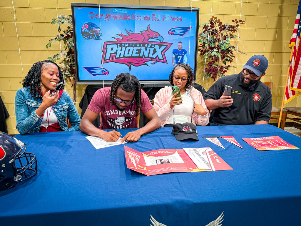 Four people seated around a table sign papers. One holds a football helmet. Banners, a flag, and a screen are in the background.