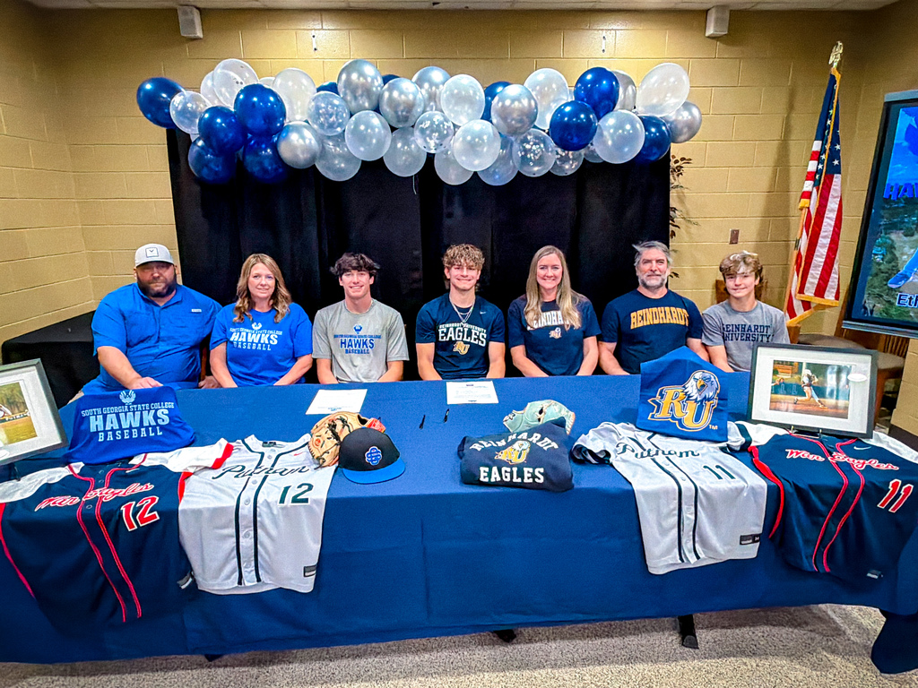 A group of people sits at a table with sports jerseys and caps. Balloons and a flag are in the background.