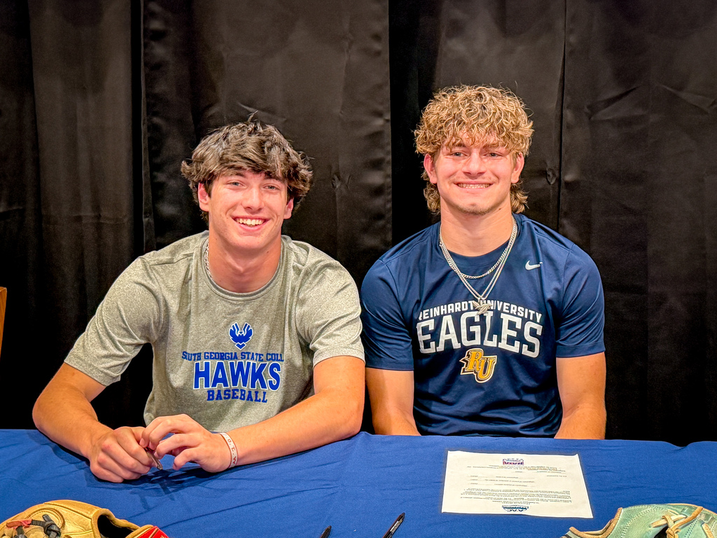 Two young men in sports jerseys seated at a table, smiling. One wears a gray shirt reading "Hawks" and the other a blue shirt reading "Eagles".