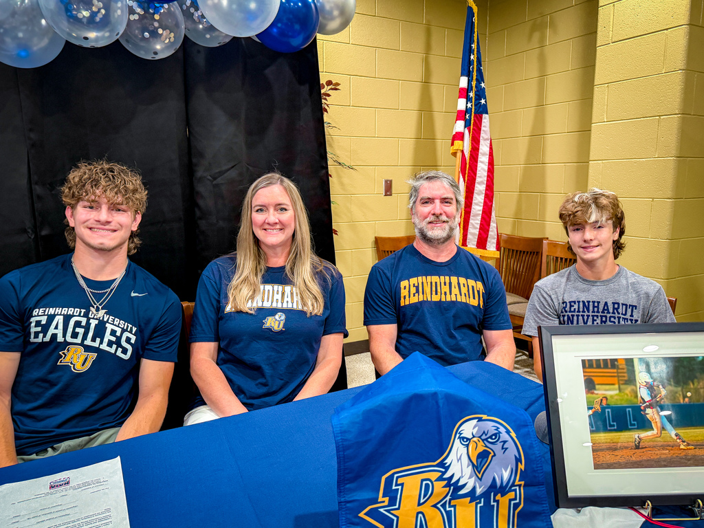 Four people wearing blue Eagles shirts smile for the camera, with a framed photo and balloons in the background.