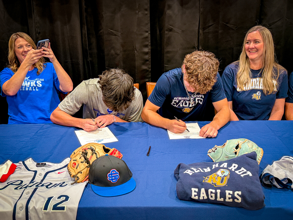 Four people sit at a table, signing documents with others nearby taking photos. Jerseys, hats, and gloves are on the table.