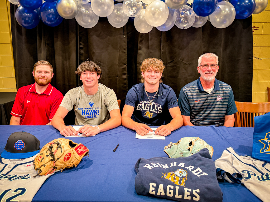 Four men sit at a table, signing papers. One wears a necklace, two wear Eagles jerseys. A baseball glove and cap are on the table.