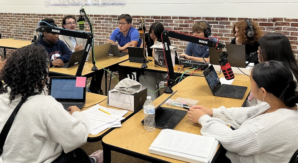 People seated at desks in a classroom with laptops, headphones, and a microphone setup.