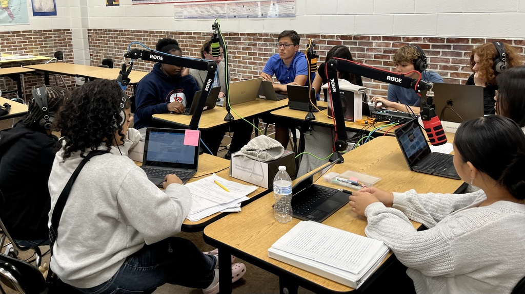 A group of students sit at desks, working on laptops, with a brick wall in the background.