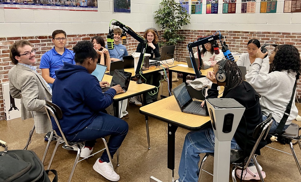 A group of students with laptops on desks in a room with brick walls. They are wearing casual clothes.