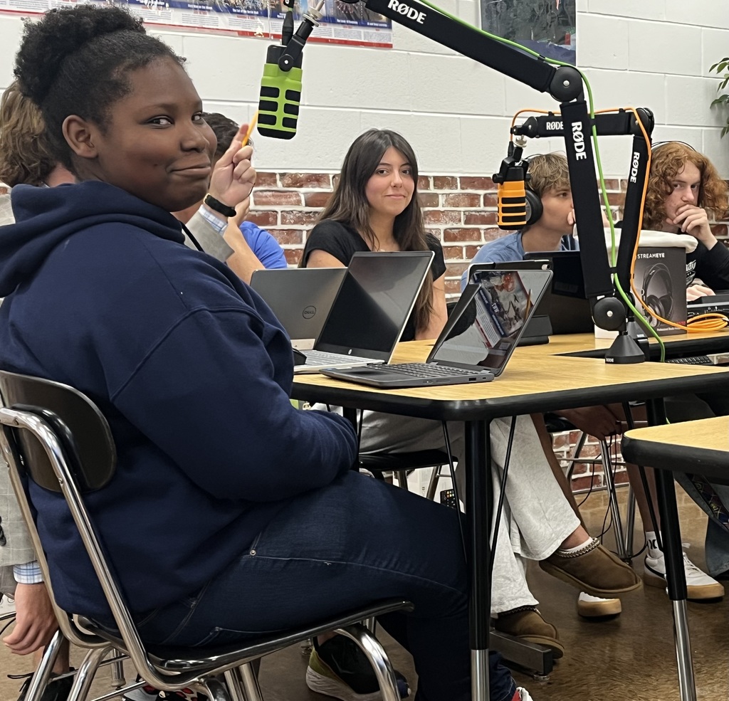Students seated around a desk, laptops open, one with a microphone, against a brick wall for podcast