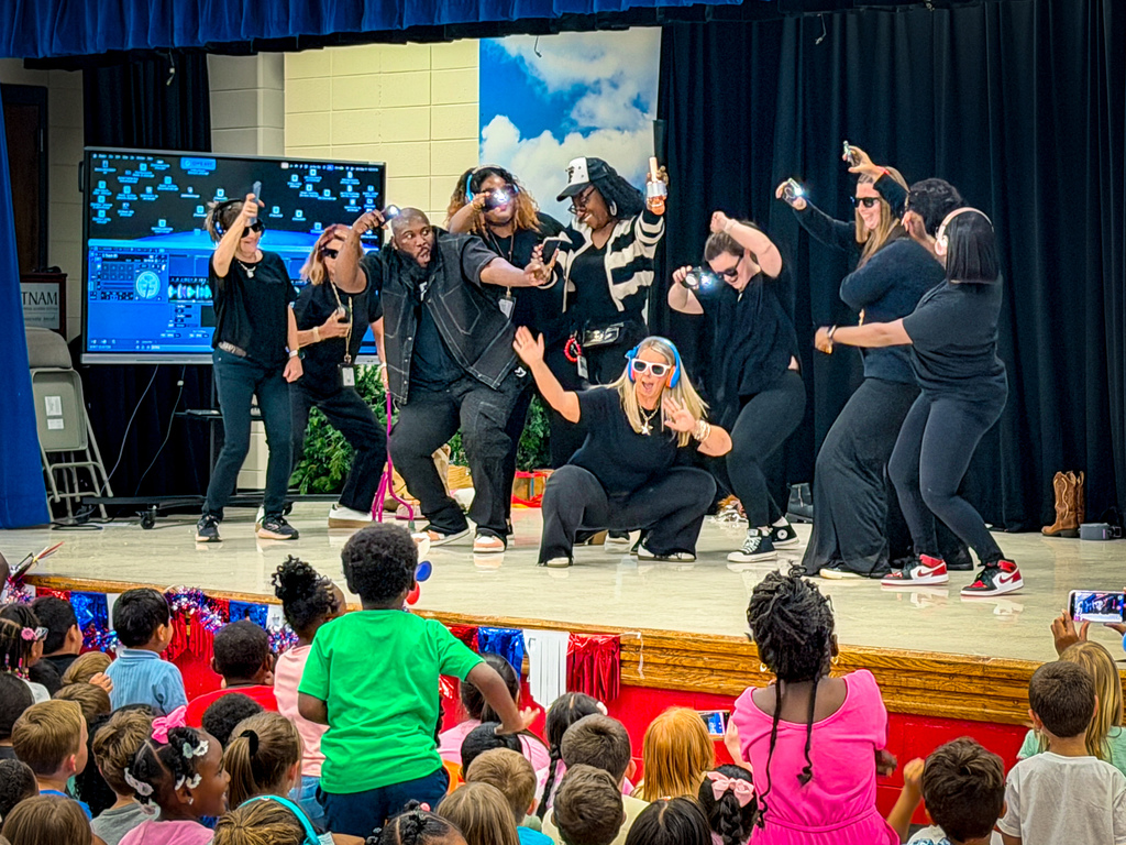 A group of adults perform dance moves on a stage with a screen and curtains in the background.