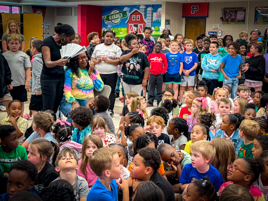 A group of children and adults gathered indoors, a woman standing at the front holding a book.
