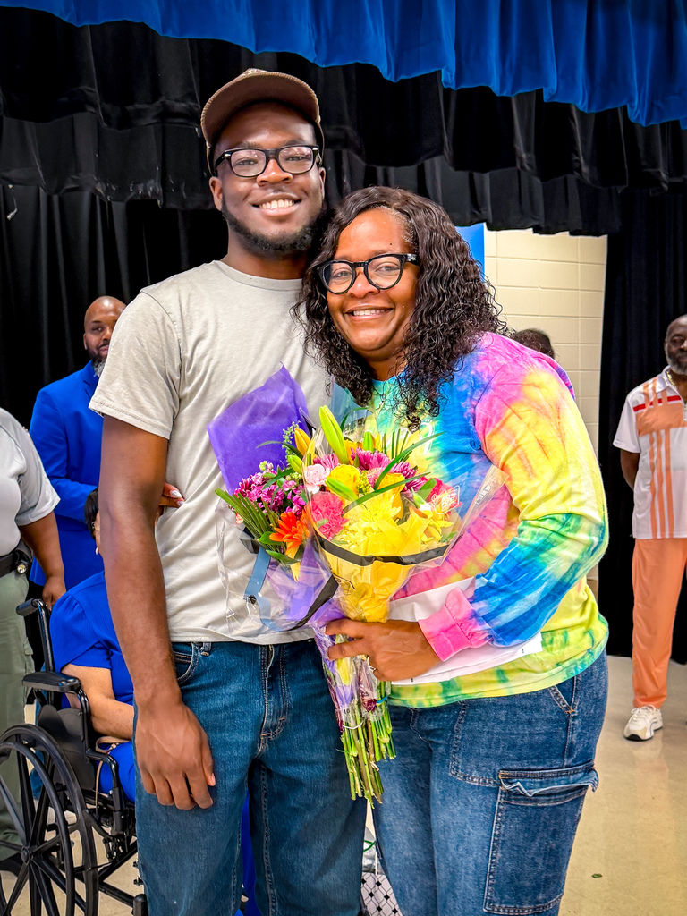 A man and woman stand smiling with a bouquet of flowers. They are surrounded by others, one in a wheelchair.