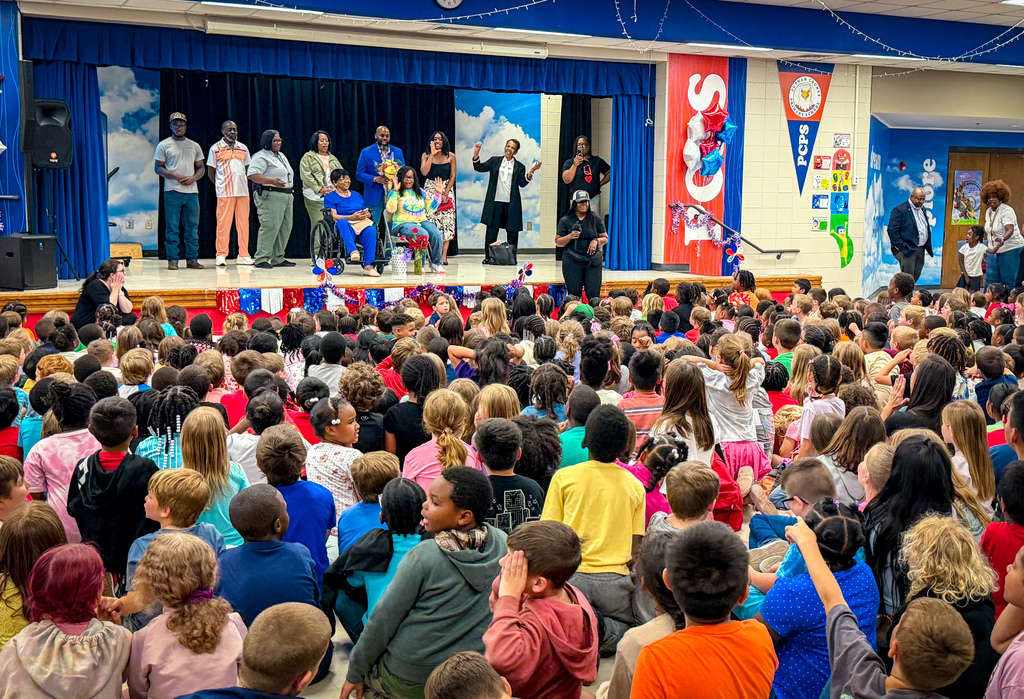 A large group of children sit in a room, watching a performance on a stage with people.