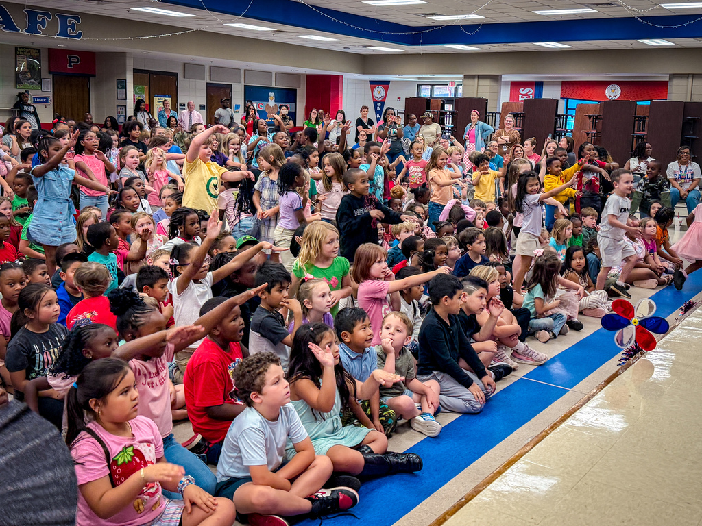A large group of children sitting and standing in rows, facing a stage in a school auditorium.