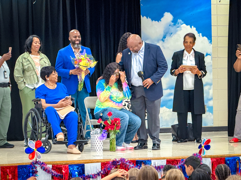 People on stage; one person in wheelchair, two holding flowers, another holding bouquet. Behind, blue sky with clouds.