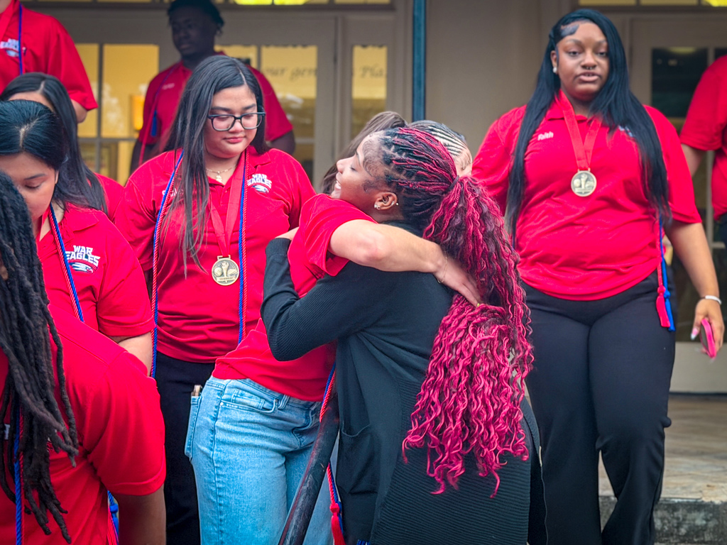 Group of people in red shirts, one hugging another person wearing a black top and curly red hair.