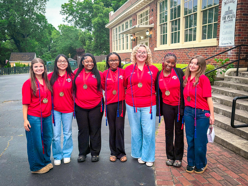 Seven women in red shirts and jeans pose in front of a brick building with stairs.