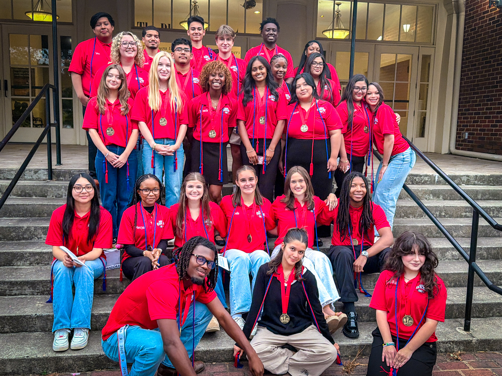 A group of people wearing red shirts and medals sit or stand on steps in front of a building.