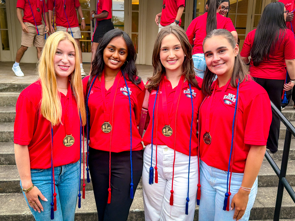 Four women in matching red shirts and sashes pose for a photo on steps with other students in red.