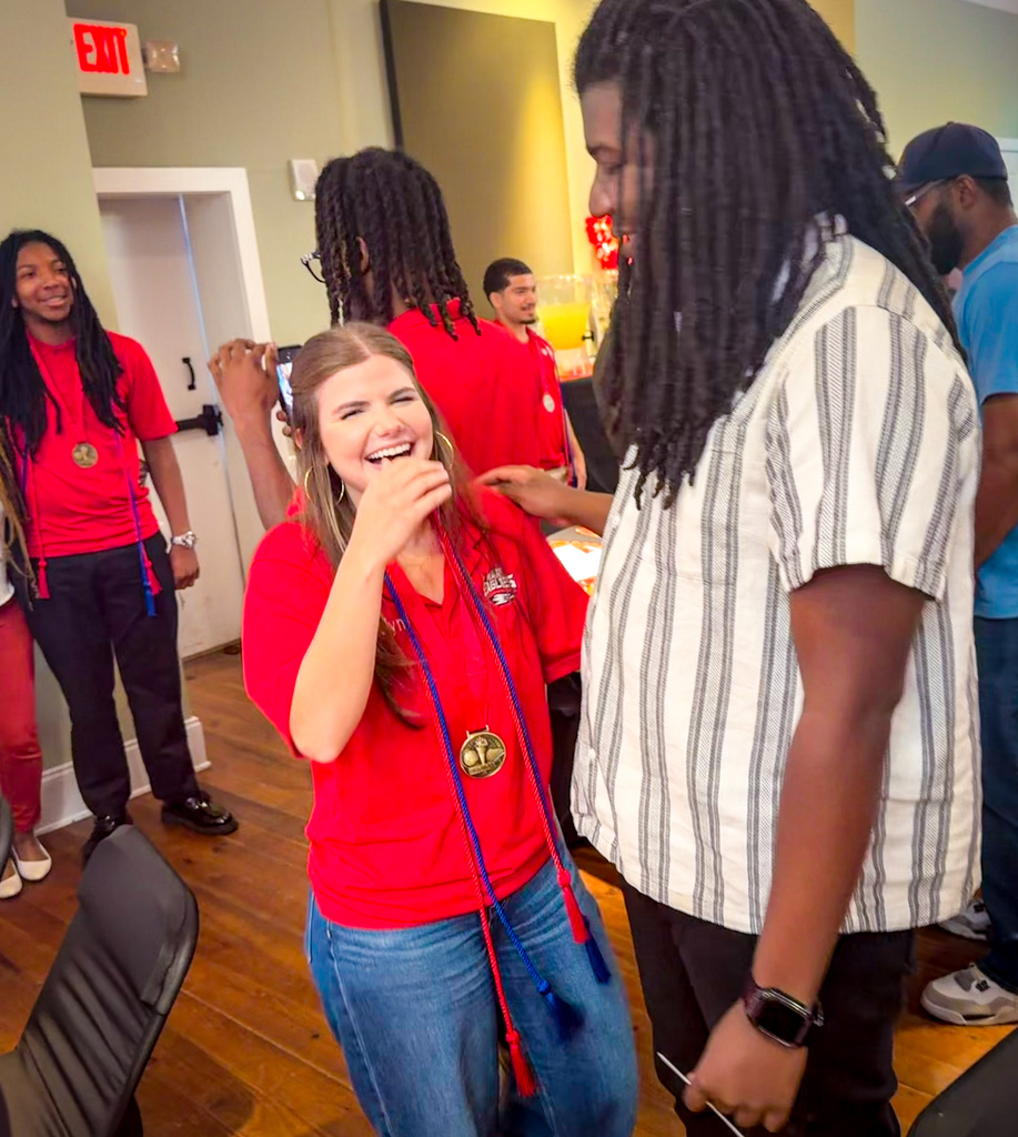 Several people in red shirts stand in a room, one of them holding a medal. A woman smiles widely.