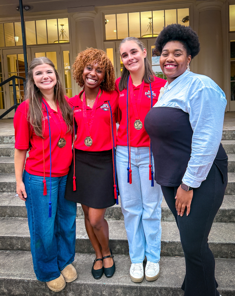 Five women in red shirts and medals stand on stairs, smiling at the camera. The woman on the far left has a ponytail.