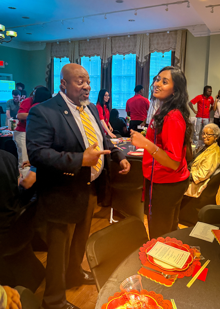A man and a woman in red shirts and ties converse at an event. Behind them, people sit or stand around tables.