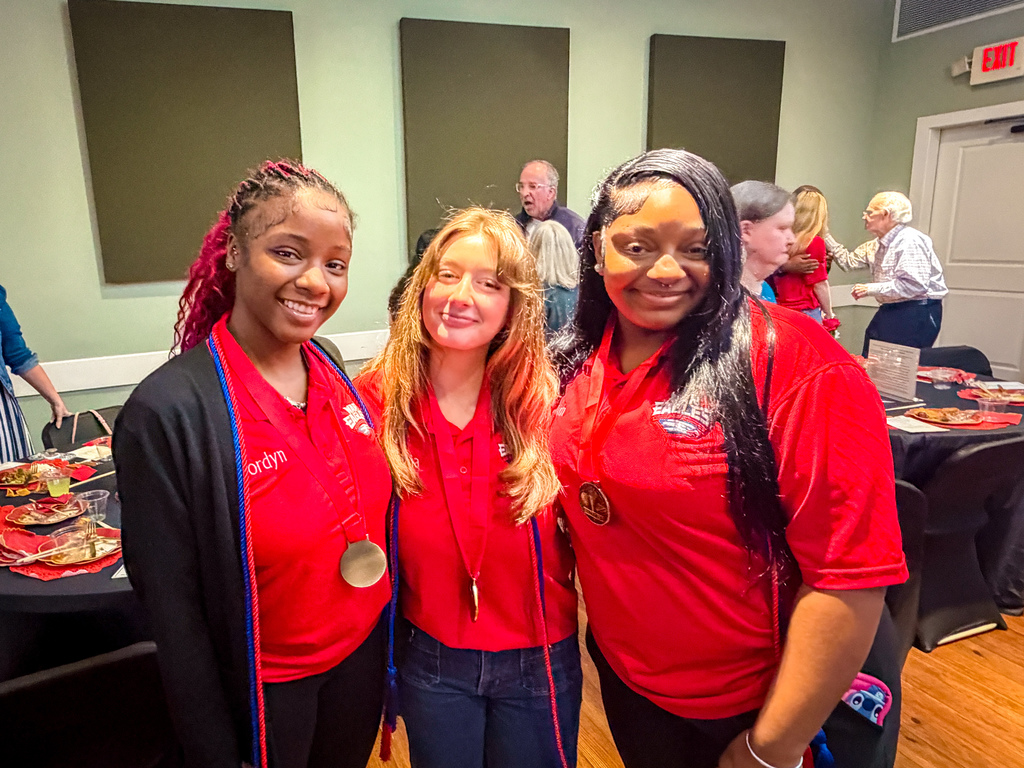 Three women in red shirts, two with medals, stand in a room with a table and others in background.