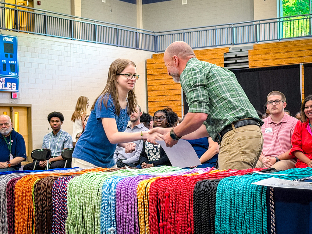 A person awards a woman a certificate at a ceremony with a large table of colorful ropes in the foreground.
