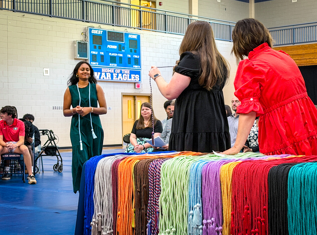 Two women stand by a table with colorful necklaces on a blue floor in a gym.