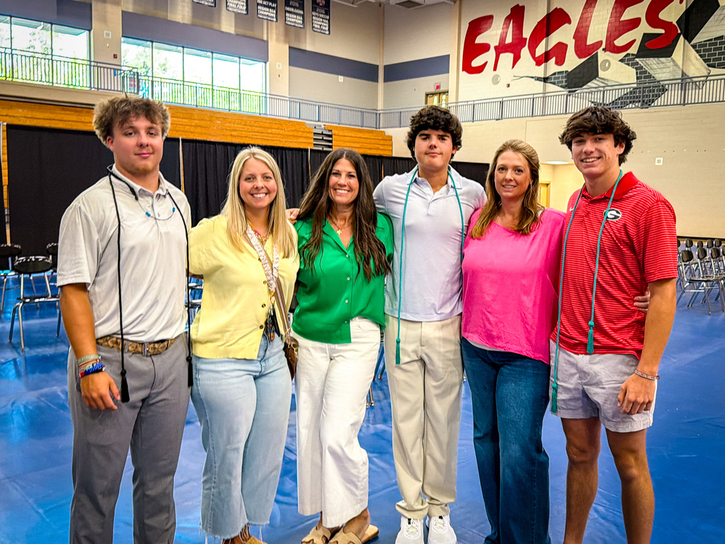 Six people stand together in a room. Five are adults and one is a young man. A sign reads "Eagles" in the background.