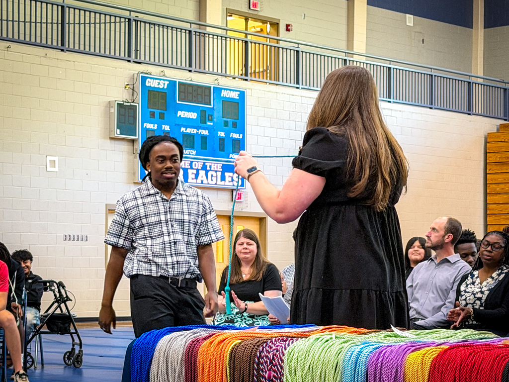 A person receiving an award at an indoor event. Audience seated, a stage with colorful fabric, a scoreboard.
