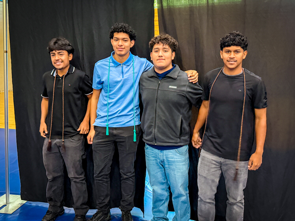 Four young men stand in a row, smiling, against a black curtain. Their casual clothing varies from black and blue to white.