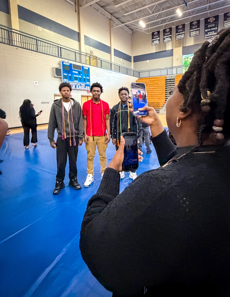 A woman captures a group of four men in a gym on a phone. One person walks in the background.