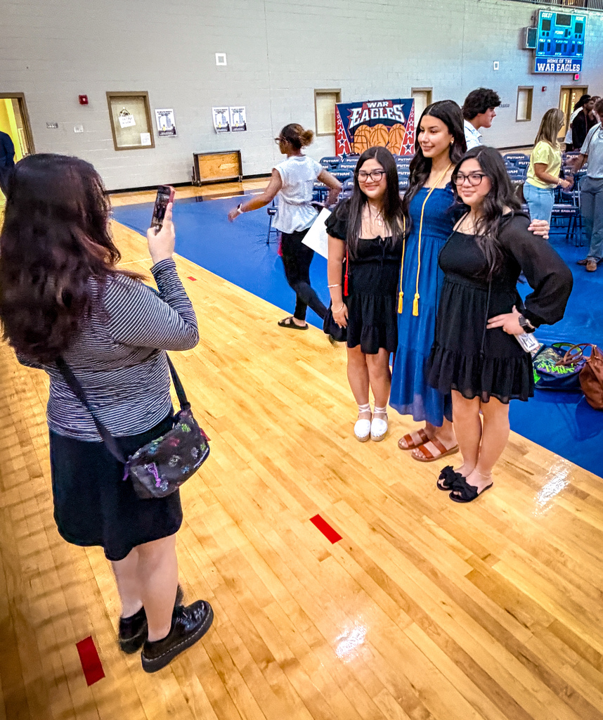 Four women, one taking a photo, in a gym with blue flooring and other people in the background.