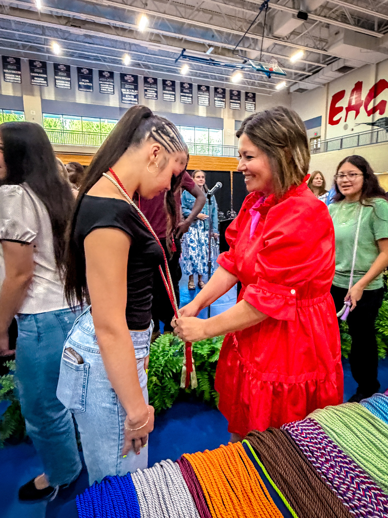 A woman in a red dress hands a necklace to a woman in blue jeans. Other people stand nearby.
